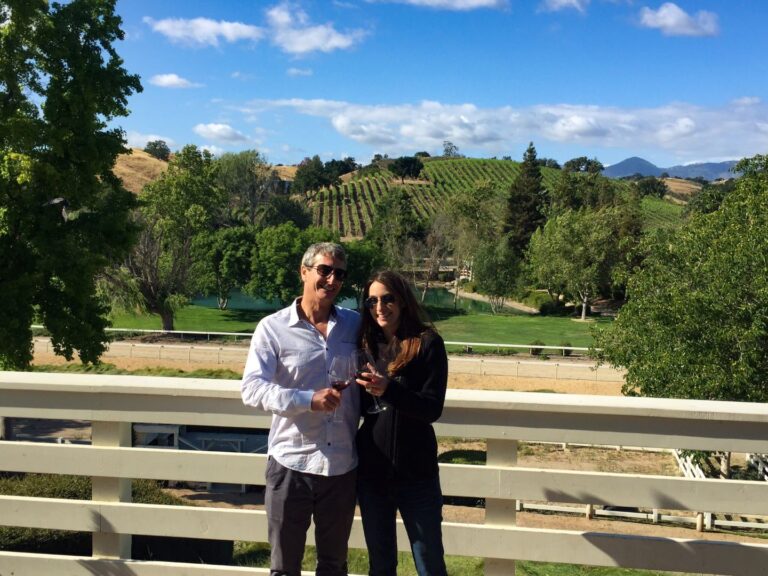A couple enjoying a wine tasting outdoors at a boutique Los Olivos winery, surrounded by oak trees and vineyard views.