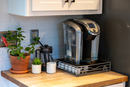 Single-serve coffee maker set up on a clean countertop with French press, mugs, and small plants for a cozy coffee station feel.