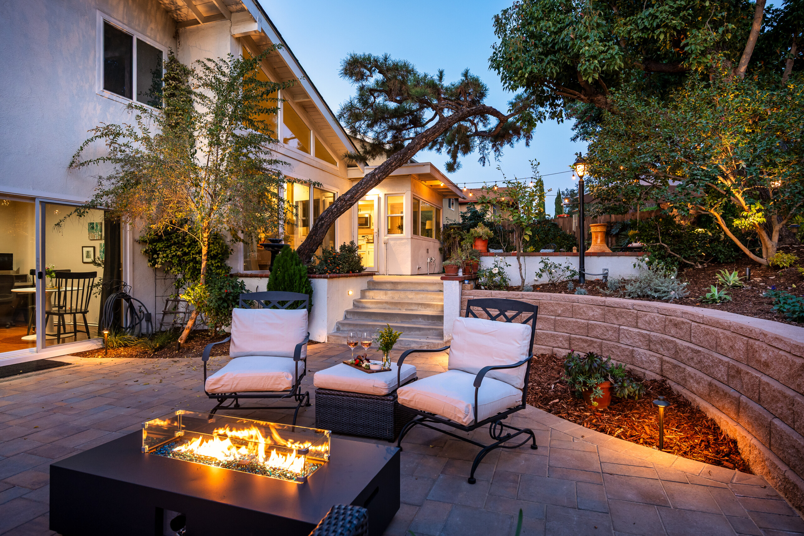Inviting backyard lounge with fire feature and ambient lighting, an ideal space to unwind in the evening, entertain guests, or enjoy peaceful outdoor moments surrounded by greenery.
